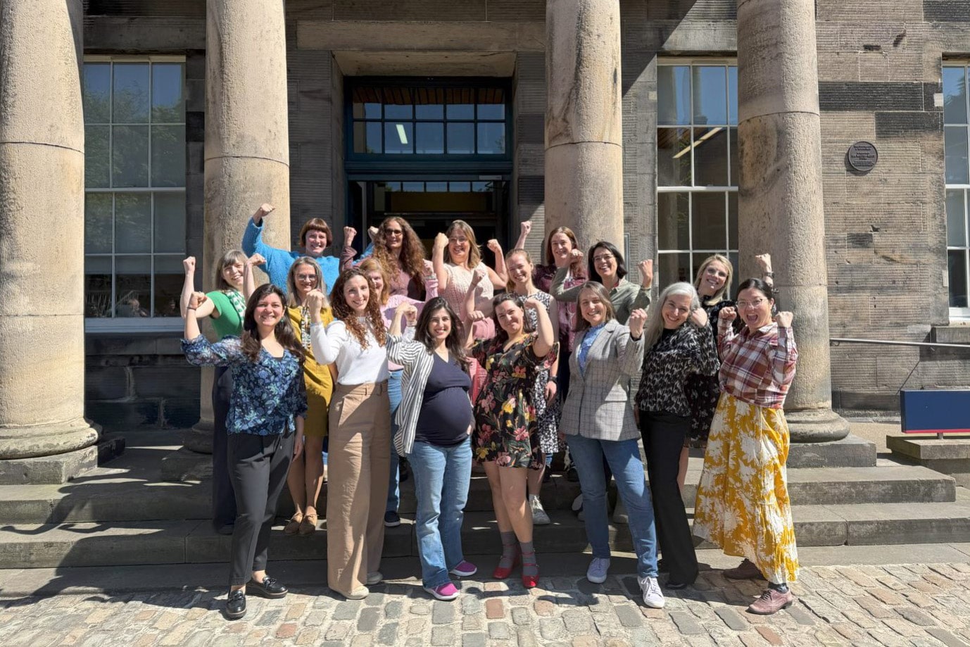 A group of academics from the Failure Modes of Engineering interdisciplinary research network stand confidently in front of a University of Edinburgh building, gesturing a show of strength and solidarity.