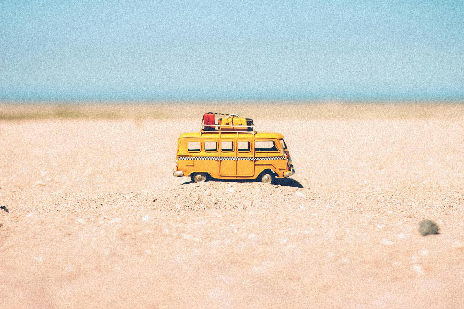 A small toy mini-bus is placed on a soft sandy beach, with a few scattered shells and a clear blue sky in the background.