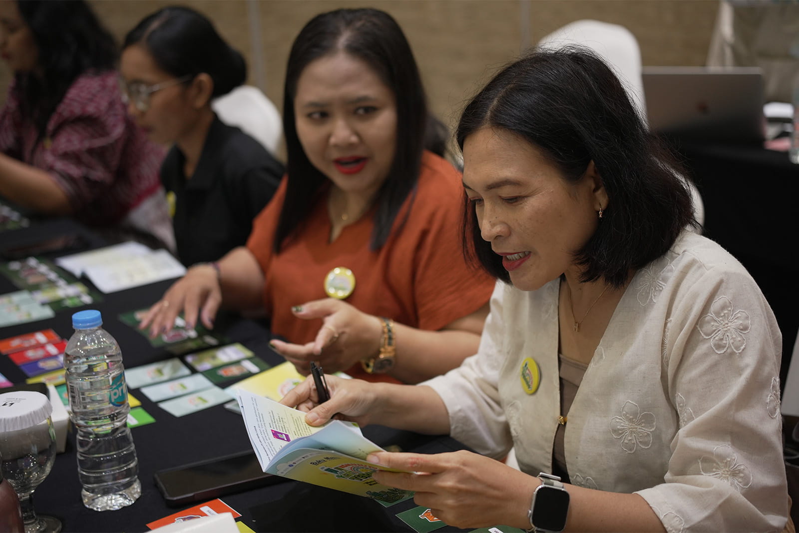 Two participants reviewing a game rulebook and cards during a purposeful play workshop session, with other attendees visible in the background.