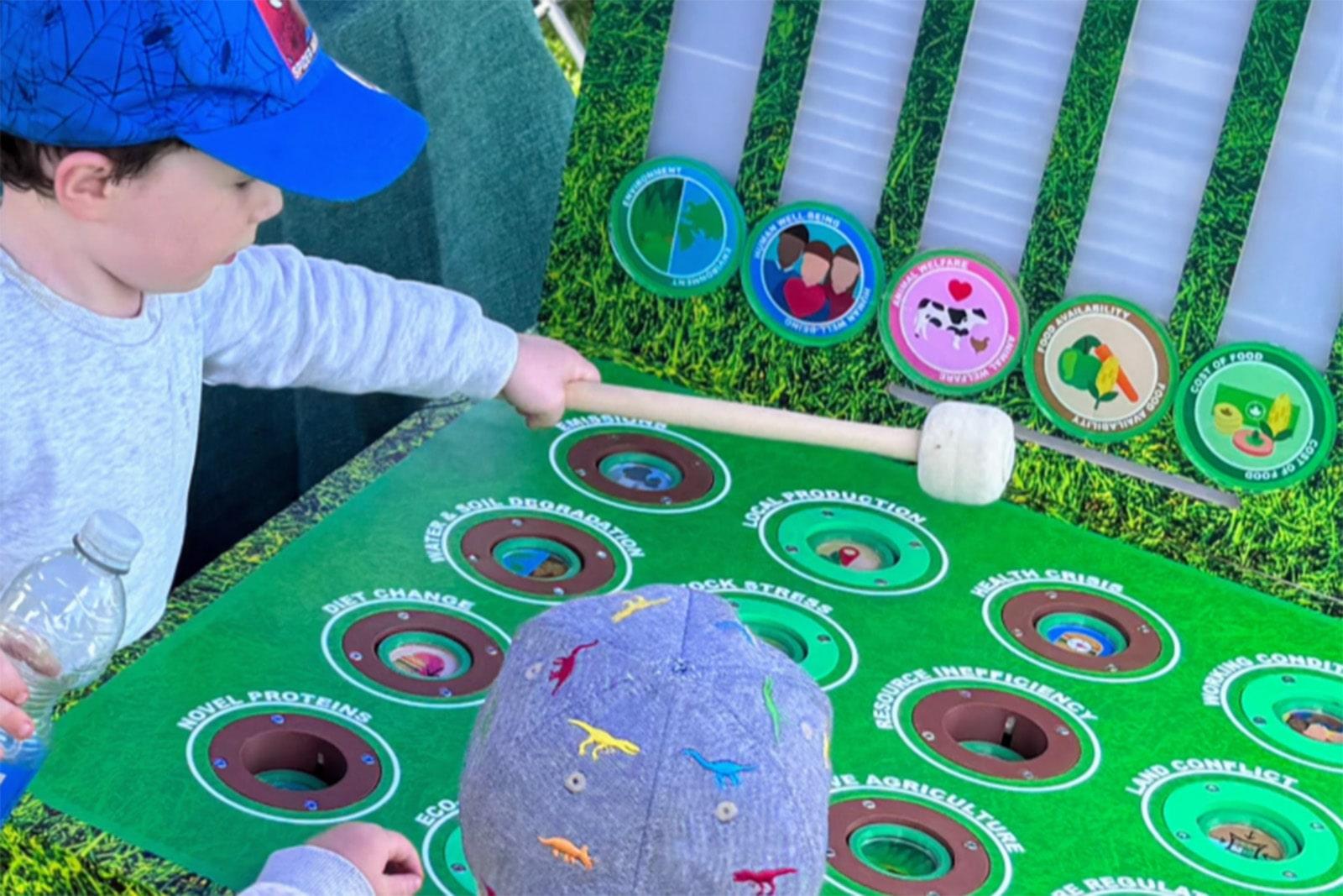 Two young boys excitedly playing a food systems-themed whac-a-mole game, hitting targets with mallets.